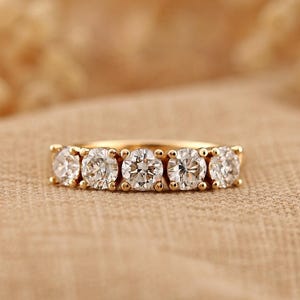 Peut inclure: Une bague en or ornée de cinq diamants ronds. Les diamants sont sertis côte à côte, créant un éclat continu. La bague est posée sur un tissu de couleur neutre.
