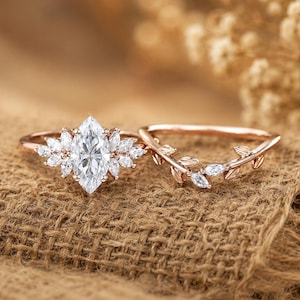 May include: A rose gold ring set consisting of two pieces. The primary ring features a central marquise-cut stone surrounded by smaller marquise-cut stones. The second ring is a curved band adorned with small metal leaves and tiny round stones.