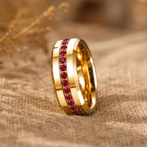 May include: A gold-toned band ring featuring a central channel set with a single row of small, round, vibrant red stones. The ring has a polished, reflective metallic finish with beveled edges, showcasing a consistent circular design throughout.