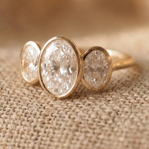 Peut inclure: Une bague en or sertie de trois pierres ovales transparentes dans un style clos. La pierre centrale est plus grande que les deux pierres latérales. L'anneau en métal est lisse et poli, présentant une teinte dorée chaude sur toute la monture.