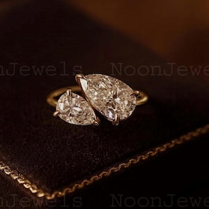 Puede incluir: Un anillo de oro con dos diamantes en forma de pera. Los diamantes están engastados uno al lado del otro, creando un diseño único y elegante. El anillo se muestra sobre una superficie marrón oscuro, con el nombre de la marca "NoonJewels" visible.