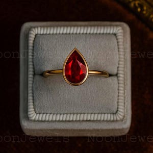 May include: A gold ring with a pear-shaped, faceted, deep red gemstone. The ring is set in a gold bezel and sits in a gray velvet ring box. The ring is displayed against a dark, textured background.
