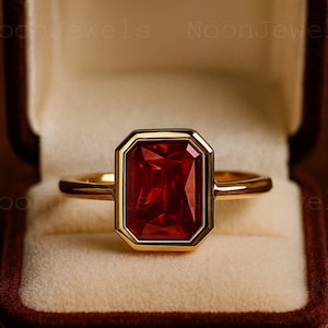 May include: A gold ring with a rectangular, deep red gemstone. The gemstone is set within a gold bezel, and the ring is displayed in a maroon velvet jewelry box. The ring's band is a simple, polished gold.
