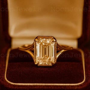 May include: An emerald-cut diamond ring in a gold band, presented in a burgundy velvet box. The ring has a rectangular diamond with a gold bezel and detailed band. The ring is displayed in a burgundy velvet box.