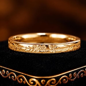 May include: A polished gold band ring featuring a delicate, continuous engraved floral pattern around the exterior surface. The ring has a smooth, reflective finish and is displayed horizontally, showcasing the intricate botanical details etched into the metal.