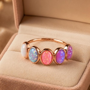 May include: A rose gold ring featuring five oval-shaped cabochon stones arranged in a row. The stones display a gradient of colours, transitioning from white to light blue, bright pink, magenta, and purple, set within a thin metallic band.