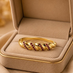 Puede incluir: Una pulsera dorada con un diseño trenzado, engastada con pequeñas piedras preciosas rojas. La pulsera se muestra dentro de un joyero beige con un interior suave y aterciopelado. El fondo es de un tono cálido y neutro.
