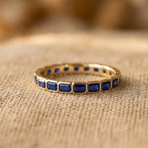 May include: A gold band ring featuring a continuous row of rectangular-cut blue sapphires set in a bezel setting. The ring is constructed with a polished gold metal frame that surrounds each individual gemstone, creating a uniform and repeating pattern.