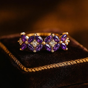 Op de afbeelding: Een gouden ring met een herhalend patroon van paarse amethiststenen in marquise-slijpvorm, gerangschikt in clusters. Tussen de paarse steengroepen bevinden zich kleine, ronde, helderwitte steentjes die in goud zijn gezet.