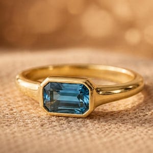 Puede incluir: Un anillo de oro con una piedra preciosa azul de corte esmeralda rectangular, montada en un engaste de bisel liso y pulido. La banda está hecha de metal dorado sólido y reflectante, con un diseño limpio que resalta la piedra azul intensa.