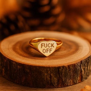 May include: Gold-toned ring with a heart-shaped face. The face is white with the words "FUCK OFF" in black block letters. The ring is set on a wooden surface with a blurred background of pine cones and leaves.
