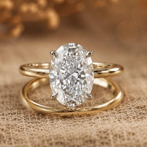 May include: A gold ring set featuring a large, clear, oval-cut center stone held by four prongs. The design consists of two smooth, polished gold bands that are joined together, creating a layered appearance with a prominent, faceted gemstone.