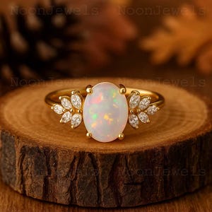 May include: A gold ring featuring a large, oval opal gemstone with iridescent colors. The ring is accented with small, clear gemstones arranged in a floral pattern on either side of the opal. The ring is set on a wooden surface.