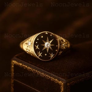 Puede incluir: Un anillo de sello dorado con una cara de esmalte negro con un diseño central de estrella con un diamante y acentos de estrella. El anillo está sobre una superficie de terciopelo marrón oscuro.