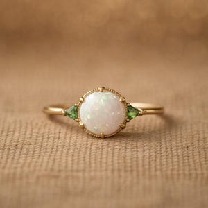 May include: A gold ring featuring a large, round, white opal center stone, flanked by two green triangular gemstones. The ring has a delicate, detailed design around the opal and a slender band.