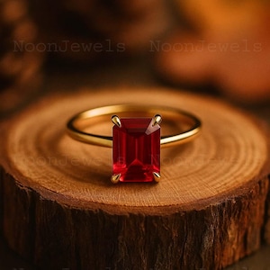 May include: A gold ring featuring a rectangular emerald-cut red gemstone held by four prongs. The ring is centered on a circular wooden slice. The text NoonJewels is visible in the background. The design includes a smooth, thin gold band.