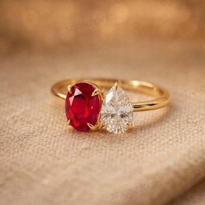 May include: A gold ring with a red oval gemstone and a pear-shaped white gemstone. The ring has a thin gold band and is set on a neutral-coloured surface.