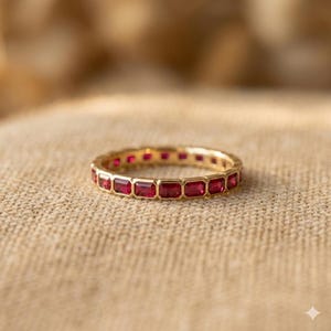 May include: A gold band ring featuring a continuous row of rectangular-cut red rubies set in a bezel style. The ring has a polished metallic finish and a uniform design that encircles the entire circumference of the band with vibrant red stones.
