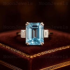Könnte beinhalten: Ein goldener Ring mit einem großen, rechteckigen, hellblauen Edelstein als Mittelstück, flankiert von zwei kleineren, rechteckigen, weißen Edelsteinen. Der Ring ist vor einem dunklen, strukturierten Hintergrund platziert.