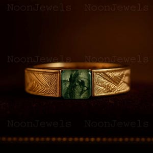 May include: A gold ring featuring a square-cut green moss agate gemstone with dark inclusions. The band is crafted from yellow gold and displays a detailed, etched leaf-like pattern on both sides of the central stone setting.