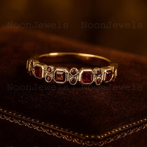 Puede incluir: Un anillo de oro con piedras preciosas rojas cuadradas y redondas alternas. El anillo está sobre una superficie marrón oscuro, con el texto "NoonJewels" visible en el fondo.