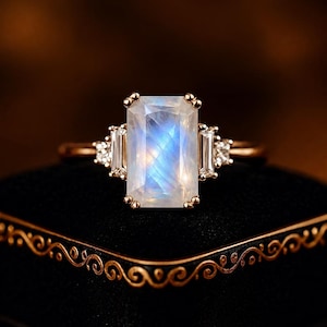 May include: A rose gold ring featuring a large rectangular moonstone centre gem with a blue iridescent sheen. The centre stone is flanked by two smaller baguette-cut side stones and tiny round accent stones set on a thin rose gold band.