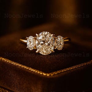 May include: A gold ring with three large, sparkling diamonds. The central oval diamond is flanked by two smaller oval diamonds. The ring is set against a dark brown velvet background, with the text "NoonJewels" visible.