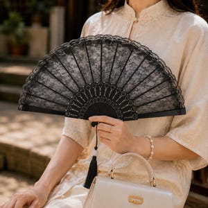 Peut inclure: Un éventail pliant noir composé d'un tissu en dentelle orné d'un motif floral, d'un bord festonné et d'une structure en plastique noir. Un pompon décoratif noir est suspendu à la base de l'éventail, qui est tenu en position centrale.