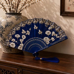 Puede incluir: Un abanico plegable de color azul intenso con intrincados bordados florales blancos y motivos de mariposas. El abanico tiene un borde festoneado tipo encaje con detalles en tono dorado y una borla azul a juego unida al mango en la base.