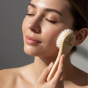 Può includere: Una spazzola per il viso in legno chiaro con setole bianche è tenuta contro la guancia di una persona. La spazzola ha un manico curvo e il testo "NATURALIZ BEAUTY" è stampato sulla testina. Gli occhi della persona sono chiusi.