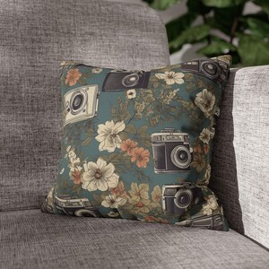 May include: A teal blue throw pillow with a floral pattern and vintage cameras. The cameras are in shades of brown and black. The flowers are in shades of white, pink, and brown.