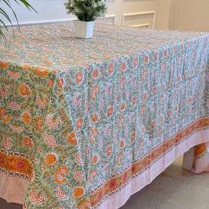 May include: A floral patterned tablecloth in shades of green, orange, and pink. The tablecloth features a repeating pattern of flowers and leaves. The border has a striped design. A small potted plant sits on the table.