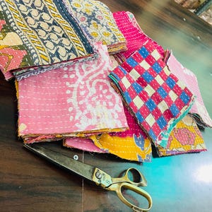 May include: A collection of colorful, patterned fabric squares in pink, blue, yellow, and white, stacked on a dark wooden surface. A pair of large, vintage brass scissors rests nearby. The fabrics feature various stitched designs.