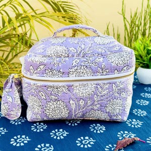 May include: A lavender cosmetic bag and small pouch with a white floral design. The bag features a top handle, a gold zip, and a quilted texture. The set is displayed on a blue patterned surface.