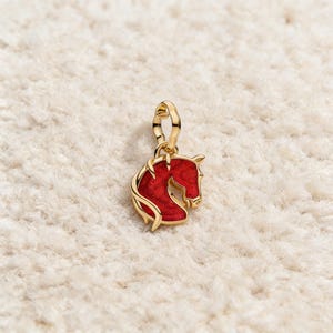 May include: A gold-coloured horse head charm with a red enamel fill. The charm is suspended from a gold-coloured bail. The horse head is a profile view, with a detailed mane and eye. The charm is set against a white, textured background.