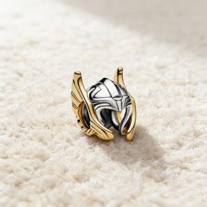 Puede incluir: Un charm dorado y plateado con forma de casco con detalles alados. El casco tiene una cara plateada y alas doradas. Este charm es probablemente un objeto de colección o una joya.
