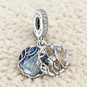 May include: Silver charm with a deer design. The charm features a deer inside a circular frame with clear gemstones. A second charm has a blue and white design. The top of the charm has the word "PANDORA" and "ALE S925".