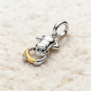 May include: A silver monkey charm with a gold-coloured banana. The charm is designed to be attached to a bracelet or necklace. The monkey's face and body are detailed, and the banana is curved.