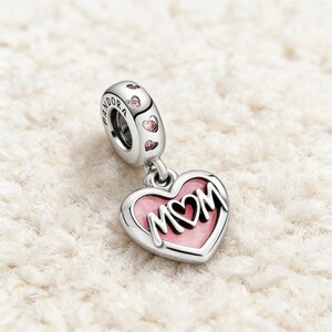 May include: A silver charm with a heart-shaped pendant that says "MOM" in white lettering on a pink background. The charm has a loop with the word "PANDORA" and pink heart accents.