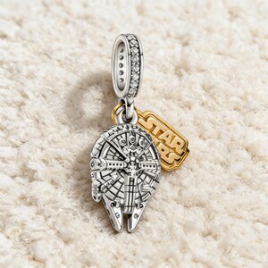 Puede incluir: Charm plateado con el diseño del Halcón Milenario y una etiqueta dorada de Star Wars. El charm tiene una superficie detallada y texturizada, y un lazo adornado con pequeñas piedras transparentes.