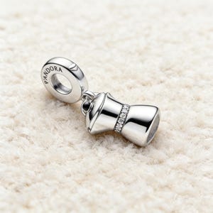 May include: A silver charm in the shape of a coffee maker. The charm features a ring with "PANDORA" inscribed. A band of small, clear stones encircles the coffee maker. The charm hangs against a white, textured backdrop.
