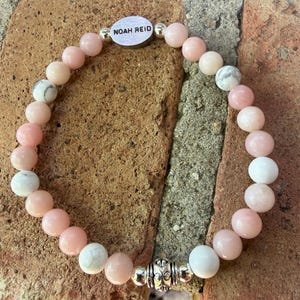 May include: A beaded bracelet featuring pink and white gemstone beads. The bracelet includes a silver-toned accent piece with the engraved text "NOAH REID". The bracelet is displayed on a textured, reddish-brown surface.