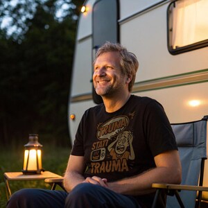 Peut inclure: Un homme porte un t-shirt noir avec le texte "TRUMA statt TRAUMA" et un dessin animé, assis dans une chaise de camping. Une lanterne allumée est posée sur une petite table. Une caravane est en arrière-plan.