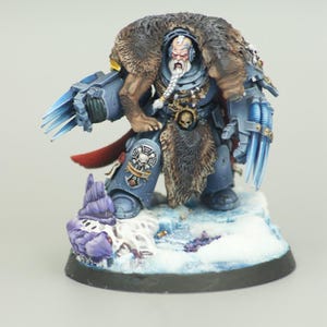 May include: A miniature figurine of a warrior in blue armor, adorned with a fur cloak and a white beard. The figure is posed on a snowy base, wielding blue energy claws. The miniature is painted with detailed shading and highlights.