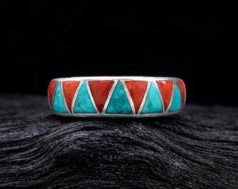 Könnte beinhalten: Ein silberner Ring mit einem geometrischen Muster aus roten und türkisfarbenen Dreiecken. Der Ring ist vor einem dunklen, strukturierten Hintergrund platziert, der die lebendigen Farben und das komplizierte Design hervorhebt. Ein auffälliges Schmuckstück.
