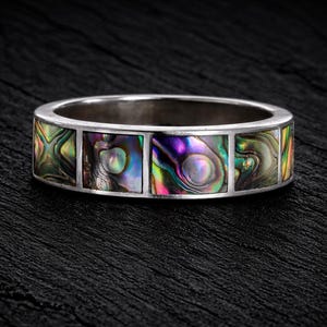 Może przedstawiać: Srebrny pierścionek z opaską inkrustowaną prostokątnymi kawałkami iryzującej muszli abalonu. Abalon prezentuje gamę kolorów, w tym zielony, fioletowy i złoty. Pierścionek jest umieszczony na ciemnym, teksturowanym tle.