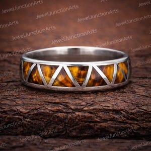 May include: A silver ring with a band featuring a geometric design of triangles and a tiger's eye stone inlay. The ring is set against a dark, textured background, showcasing the jewelry's details.