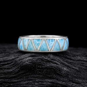 May include: A silver band ring featuring a series of inlaid triangular pieces of light blue, marbled larimar stone. The metal frame creates a geometric pattern around the circumference of the ring, showcasing the distinct sky-blue color.
