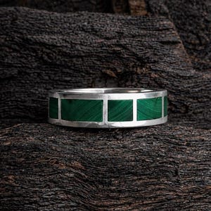 Puede incluir: Un anillo de plata con incrustaciones rectangulares de malaquita verde. El anillo se encuentra sobre una superficie de madera oscura y texturizada, resaltando el contraste entre la piedra verde brillante y la plata.
