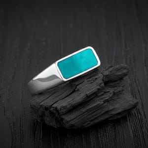 Puede incluir: Un anillo de plata con una piedra rectangular de turquesa. El anillo tiene un acabado pulido y un diseño moderno. La piedra turquesa es de un tono azul-verdoso vibrante. El anillo se muestra sobre un trozo de madera oscura y texturizada.